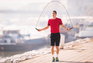 Young man wearing sportswear skipping rope at quay during autumn clipart