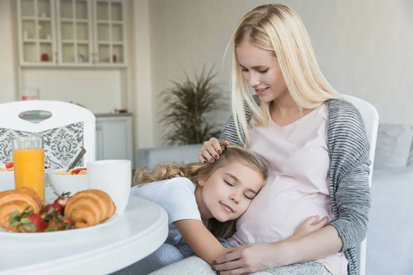 台所で妊娠中の母親の腹の上で眠っている陽気な娘 — ストック写真