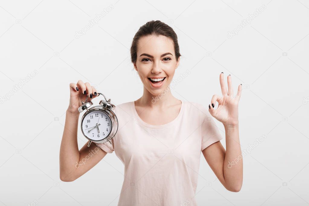 beautiful girl showing ok gesture and holding alarm clock isolated on white