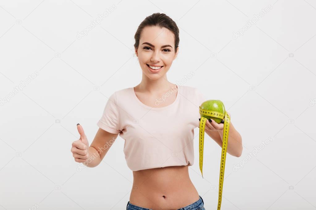 woman holding apple tied with measuring tape and showing thumb up isolated on white