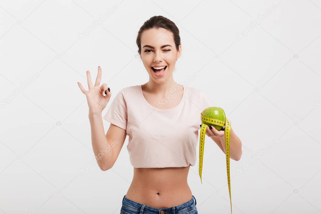 woman holding apple tied with measuring tape and showing okay sign isolated on white