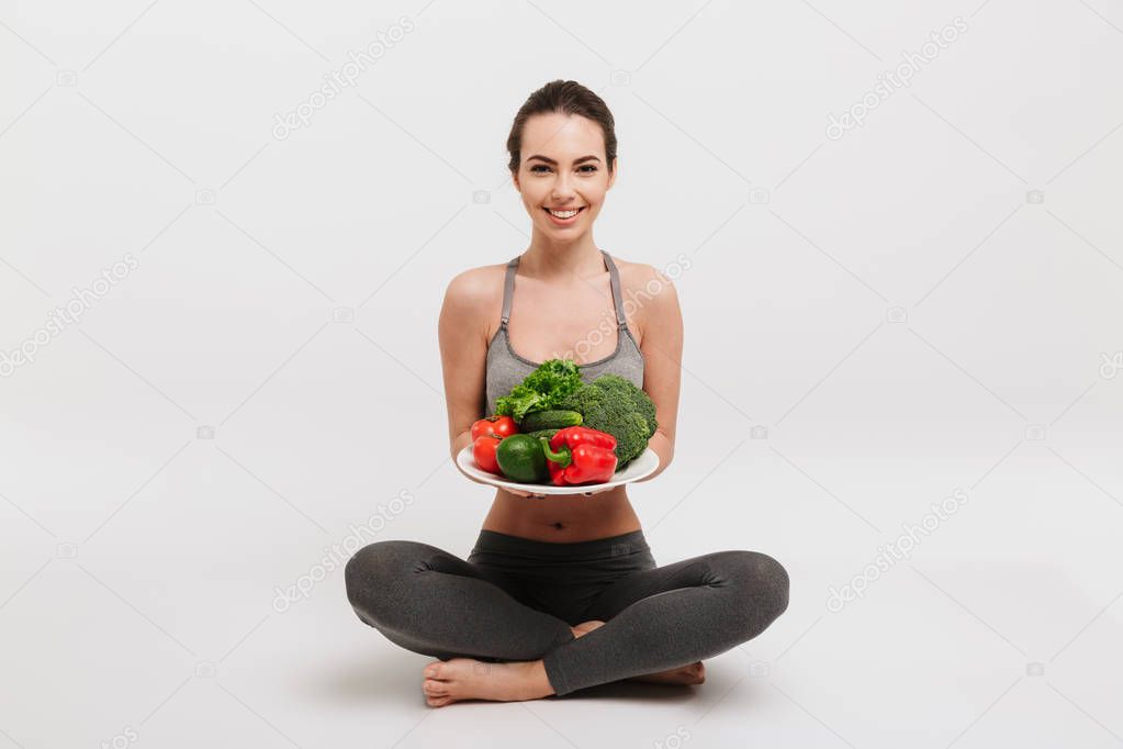 beautiful young woman sitting on floor with tray of various healthy vegetables isolated on white