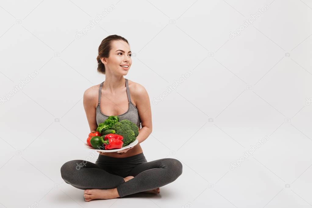 happy young woman sitting on floor with tray of various healthy vegetables isolated on white