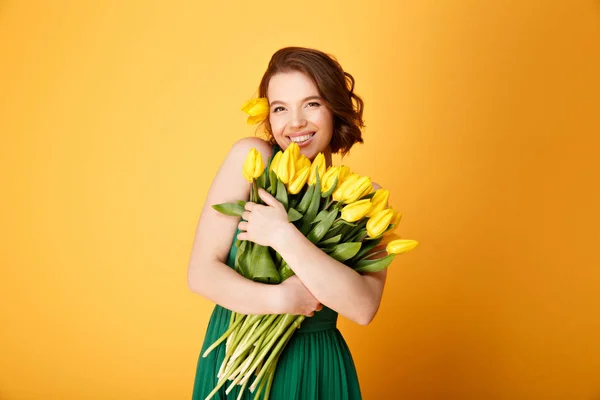 portrait of smiling woman with bouquet of yellow tulips in hands isolated on orange