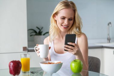 beautiful smiling blonde girl holding cup of coffee and using smartphone during breakfast clipart
