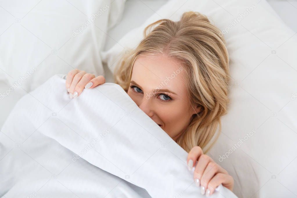 high angle view of beautiful blonde girl lying under blanket and smiling at camera