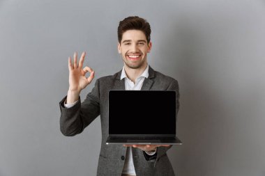 portrait of smiling businessman in suit with laptop with blank screen showing ok sign against grey wall background