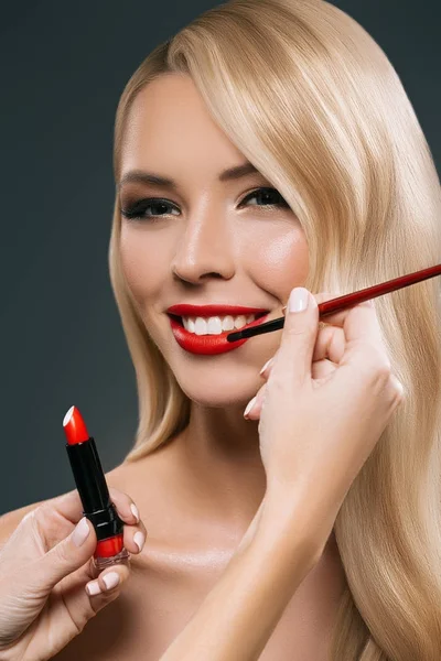 Portrait of beautiful blonde hair girl with visagiste hands applying lipstick with brush, isolated on black — Stock Photo