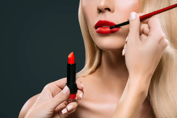 Cropped image of beautiful blonde hair girl with visagiste applying lipstick with brush, isolated on black — Stock Photo