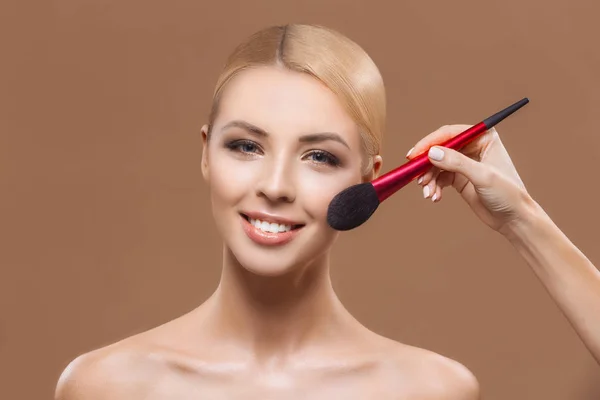 Schöne Frau, die Puder mit Schminkpinsel aufträgt, isoliert auf braun — Stockfoto