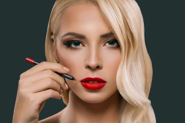 Beautiful blonde woman making red lips with cosmetic pencil, isolated on grey — Stock Photo
