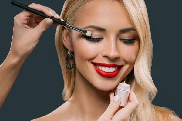 Hermosa mujer rubia aplicando maquillaje glamoroso con sombras de ojos, aislado en gris - foto de stock