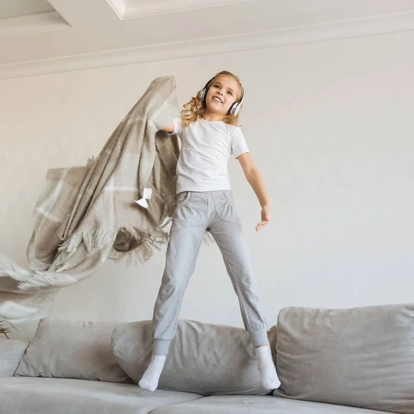 Smiling kid jumping on sofa and listening music with headphones — Stock Photo