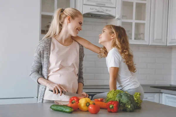 Filha abraçando mãe grávida enquanto ela cozinha na cozinha — Fotografia de Stock