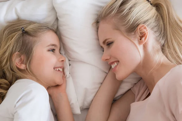 Vista superior de sonriente hija y madre acostadas en la cama y mirándose mutuamente - foto de stock