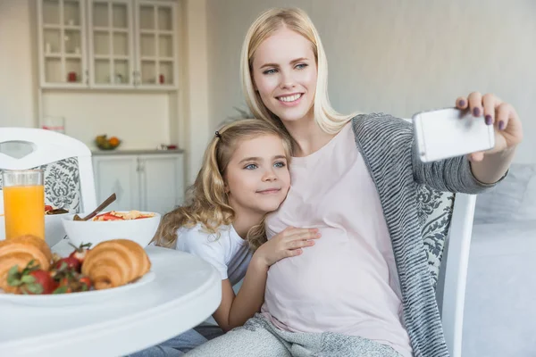 Улыбающиеся беременные мать и дочь делают селфи на кухне — стоковое фото
