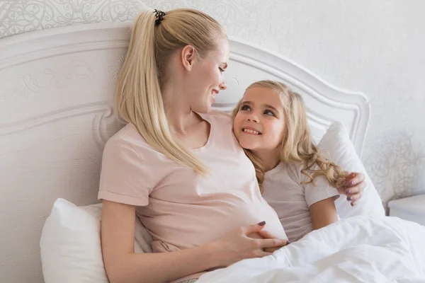 Lächelnde schwangere Mutter und Tochter liegen im Bett — Stockfoto