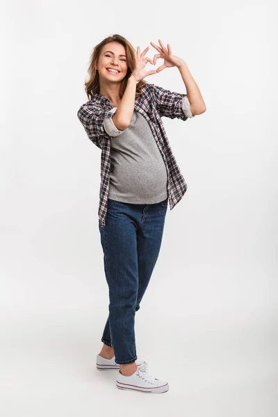 Mujer embarazada sonriente mostrando el signo del corazón aislado en gris - foto de stock