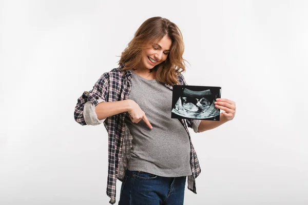 Smiling pregnant woman with ultrasound scan pointing at tummy isolated on grey — Stock Photo