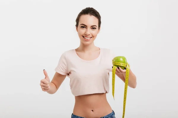 Mujer sosteniendo manzana atada con cinta métrica y mostrando el pulgar hacia arriba aislado en blanco - foto de stock