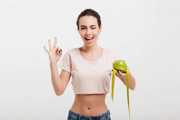 Mujer sosteniendo manzana atada con cinta métrica y mostrando signo bien aislado en blanco - foto de stock