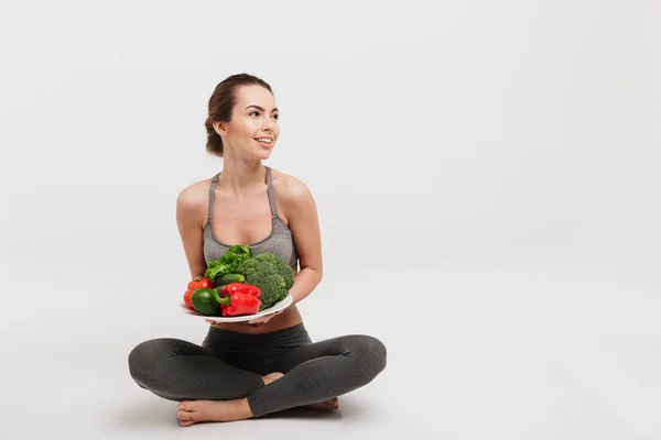 Feliz joven sentada en el suelo con bandeja de varias verduras saludables aisladas en blanco — Stock Photo