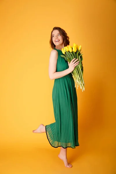 Fröhliche Frau in grünem Kleid mit Strauß gelber Tulpen isoliert auf orange — Stockfoto