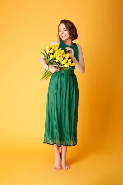 Smiling woman in green spring dress looking at bouquet of yellow tulips isolated on orange — Stock Photo