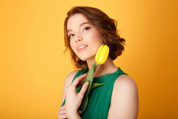 Porträt einer attraktiven Frau in grünem Kleid mit gelber Tulpe isoliert auf orange — Stockfoto