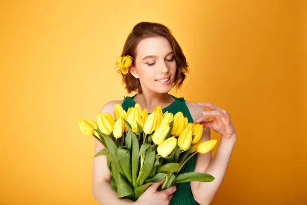 Portrait of attractive woman looking at bouquet of yellow tulips in hand isolated on orange — Stock Photo
