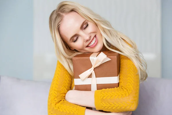 Belle fille blonde heureuse avec les yeux fermés étreignant boîte cadeau à la maison — Photo de stock