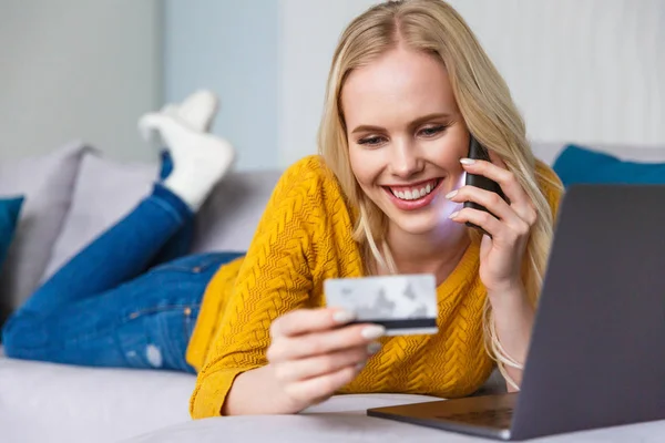 Atractiva chica rubia feliz sosteniendo la tarjeta de crédito y hablando por teléfono inteligente mientras está acostado con el ordenador portátil en casa - foto de stock