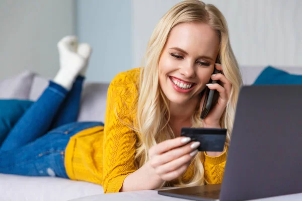 Hermosa joven sonriente sosteniendo tarjeta de crédito y hablando por teléfono inteligente en casa - foto de stock
