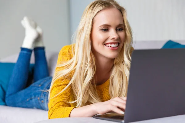 Belle fille blonde souriante couchée et utilisant un ordinateur portable à la maison — Photo de stock