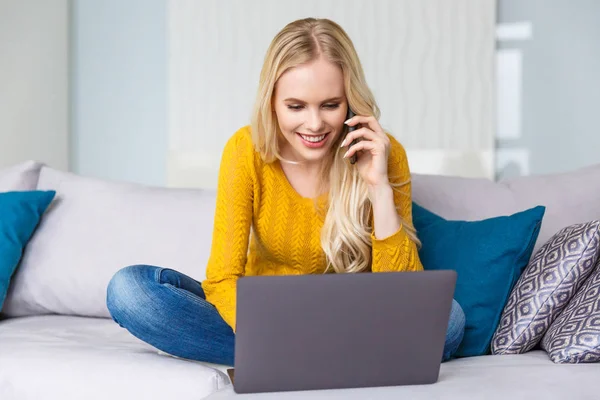 Hermosa chica rubia sonriente usando el ordenador portátil y hablando por teléfono inteligente en casa - foto de stock