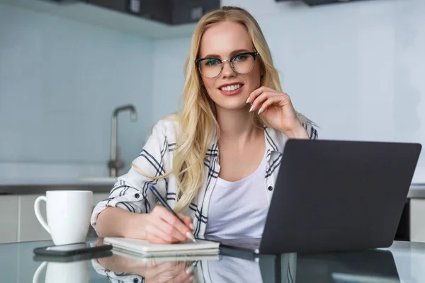 Beautiful young woman in eyeglasses smiling at camera while using laptop and taking notes at home — Stock Photo