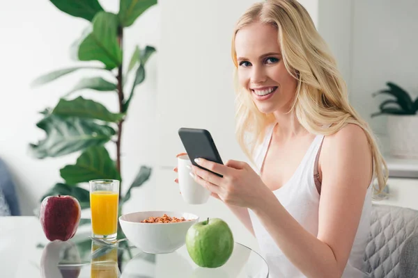 Bela jovem mulher usando smartphone e sorrindo para a câmera durante o café da manhã em casa — Fotografia de Stock