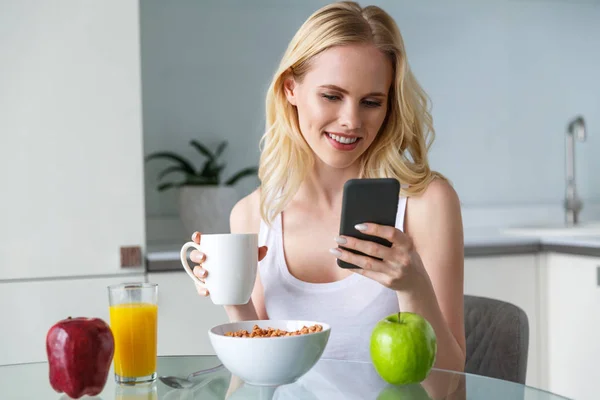 Beautiful smiling blonde girl holding cup of coffee and using smartphone during breakfast — Stock Photo