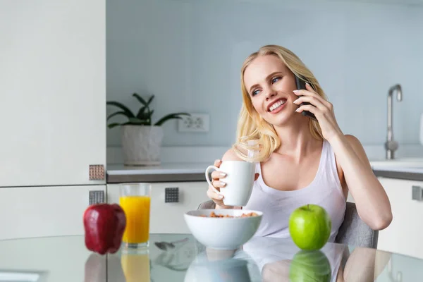 Belle jeune femme souriante boire du café et parler par smartphone pendant le petit déjeuner — Photo de stock