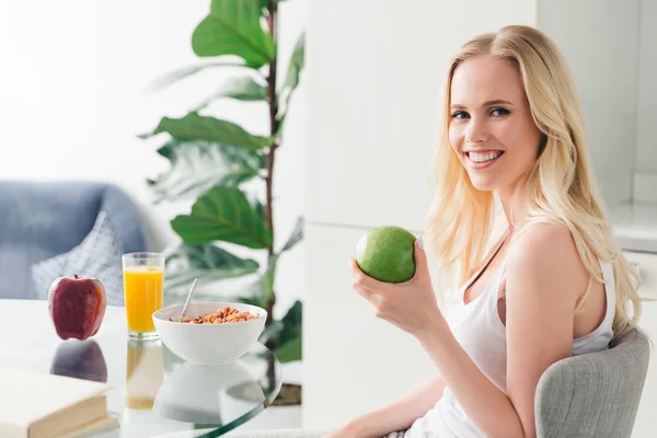 Schöne blonde Mädchen hält Apfel und lächelt in die Kamera, während sie zu Hause frühstückt — Stockfoto