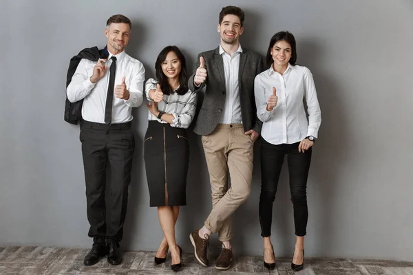 Pessoas de negócios multiculturais em desgaste formal mostrando polegares para cima enquanto em pé na parede cinza — Fotografia de Stock