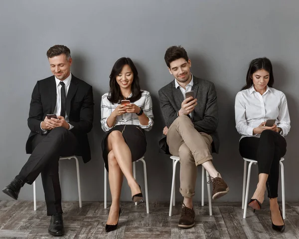 Улыбающиеся межрасовые бизнесмены в формальной одежде с помощью смартфонов в ожидании собеседования — стоковое фото