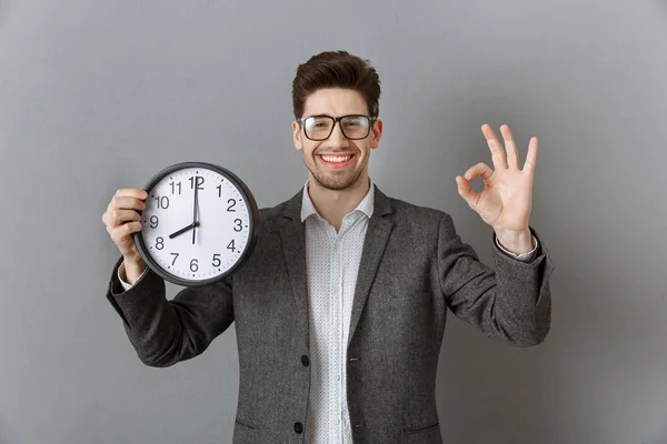 Retrato de homem de negócios sorridente com relógio na mão mostrando sinal ok no fundo da parede cinza — Fotografia de Stock
