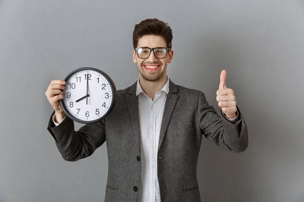 Portrait of smiling businessman with clock in hand showing thumb up on grey wall background — Stock Photo