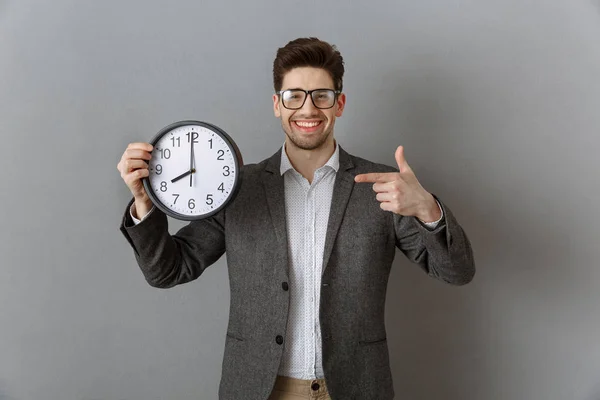 Ritratto di un uomo d'affari sorridente che punta l'orologio in mano e guarda la macchina fotografica sullo sfondo grigio della parete — Foto stock