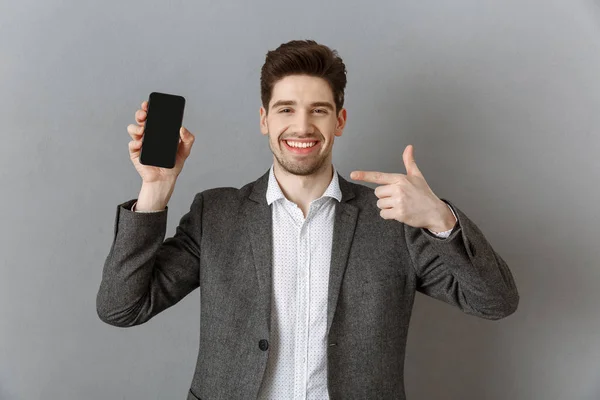 Ritratto di un uomo d'affari sorridente che punta lo smartphone con lo schermo bianco in mano sullo sfondo grigio della parete — Foto stock