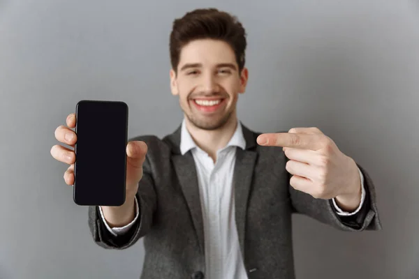 Selective focus of smiling businessman pointing at smartphone with blank screen in hand against grey wall background — Stock Photo