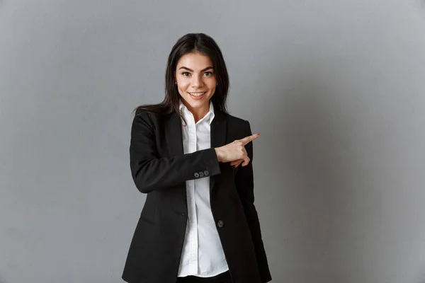 Portrait of smiling businesswoman in suit pointing away against grey wall backdrop — Stock Photo
