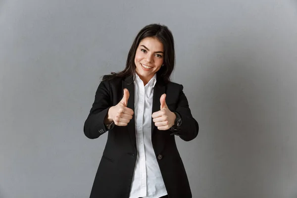 Retrato de mujer de negocios sonriente en traje mostrando pulgares hacia arriba contra fondo de pared gris - foto de stock