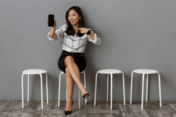 Sonriente asiático mujer de negocios apuntando a smartphone con pantalla en blanco mientras espera a entrevista de trabajo - foto de stock